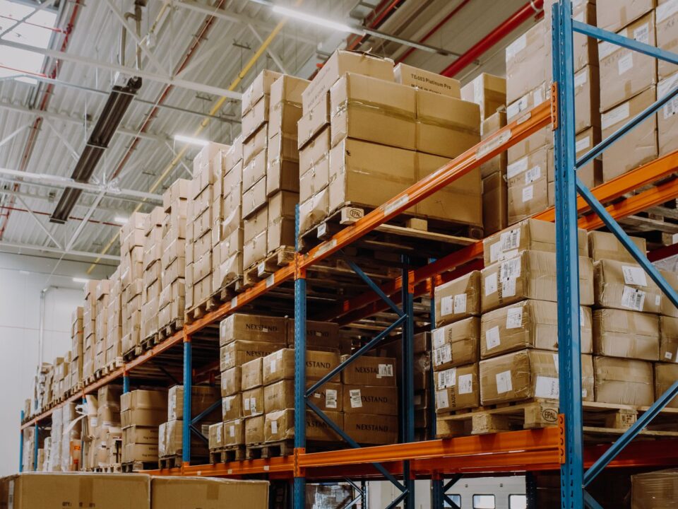 Warehouse interior with tall blue and orange shelves stacked with numerous brown cardboard boxes. Bright lighting and high ceiling create an organized atmosphere.