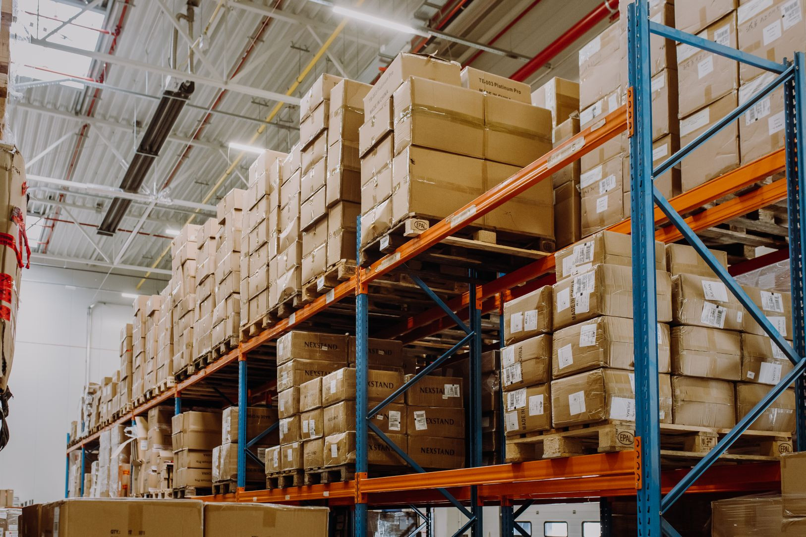 Warehouse interior with tall blue and orange shelves stacked with numerous brown cardboard boxes. Bright lighting and high ceiling create an organized atmosphere.