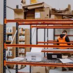A warehouse worker in an orange vest is organizing boxes on a metal shelf. Cardboard boxes and products surround him, creating a busy, industrious atmosphere.