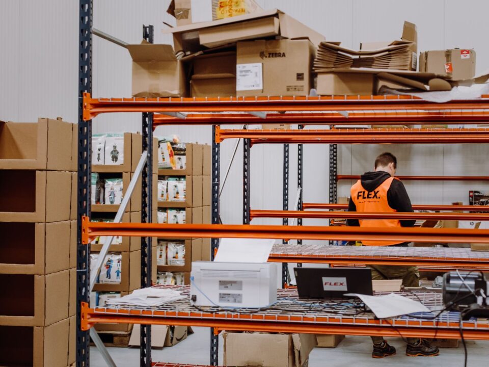 A warehouse worker in an orange vest is organizing boxes on a metal shelf. Cardboard boxes and products surround him, creating a busy, industrious atmosphere.