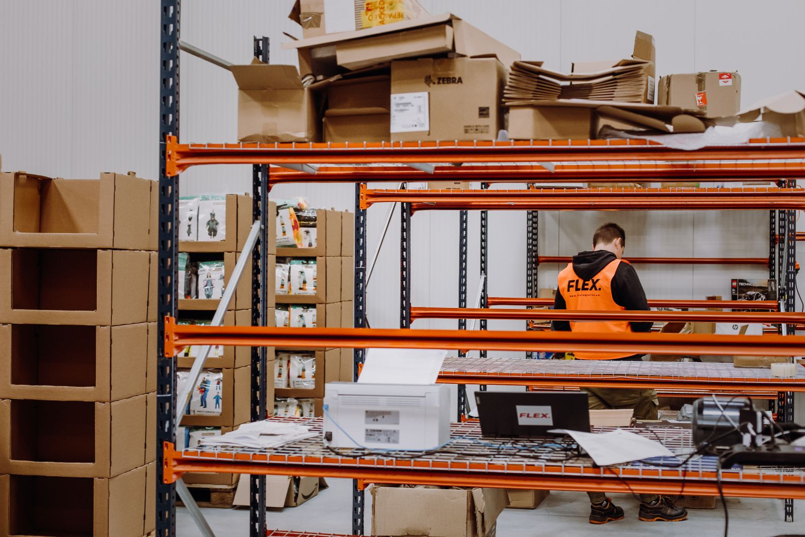 A warehouse worker in an orange vest is organizing boxes on a metal shelf. Cardboard boxes and products surround him, creating a busy, industrious atmosphere.