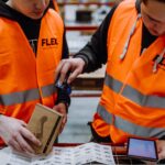 Two workers in orange safety vests use a scanner and tablet to check packages and labels in a warehouse. The scene is focused and industrious.
