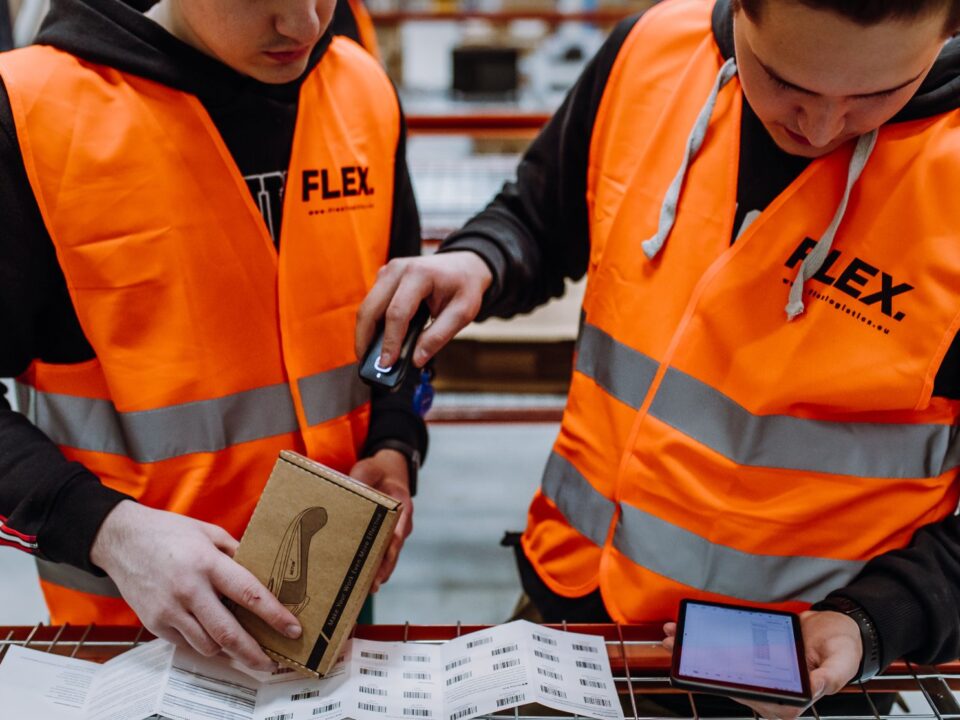 Two workers in orange safety vests use a scanner and tablet to check packages and labels in a warehouse. The scene is focused and industrious.