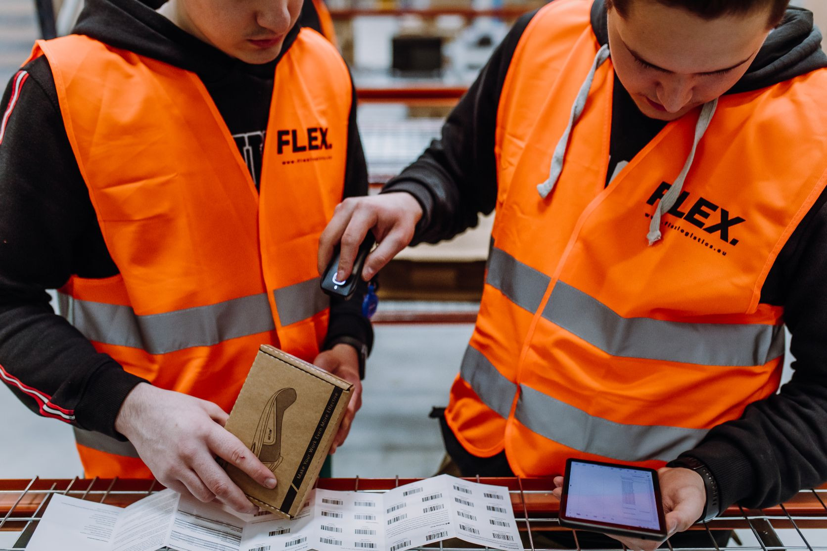 Two workers in orange safety vests use a scanner and tablet to check packages and labels in a warehouse. The scene is focused and industrious.