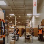 Workers in orange vests organize shelves in a spacious warehouse filled with boxes. The scene reflects orderliness and efficiency.
