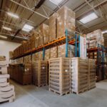 Spacious warehouse interior filled with neatly stacked cardboard boxes on shelves and pallets. Overhead lighting casts a bright, organized atmosphere.