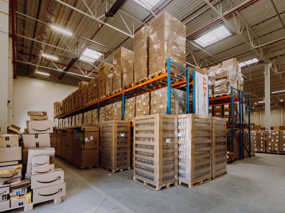 Spacious warehouse interior filled with neatly stacked cardboard boxes on shelves and pallets. Overhead lighting casts a bright, organized atmosphere.