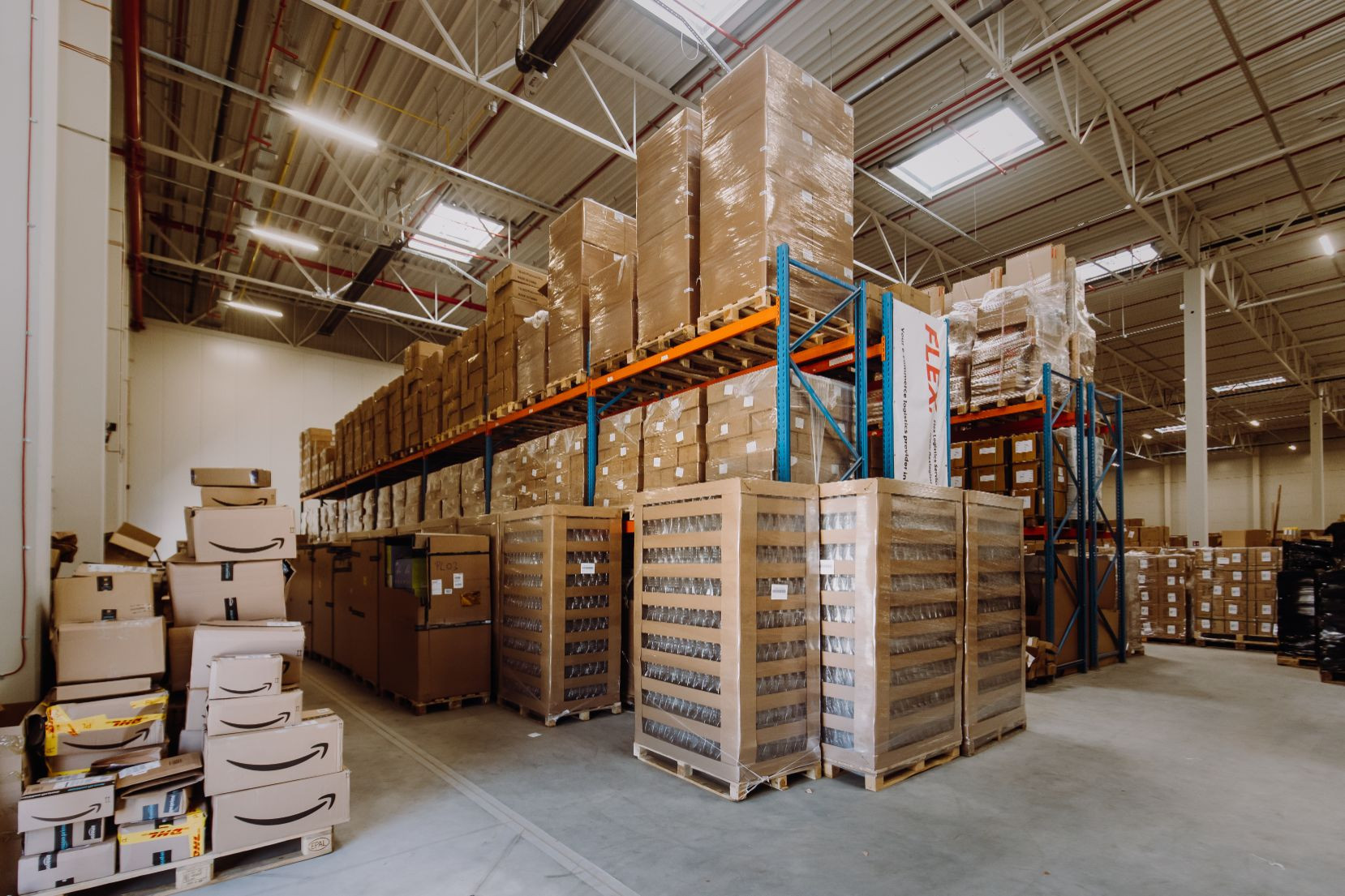 Spacious warehouse interior filled with neatly stacked cardboard boxes on shelves and pallets. Overhead lighting casts a bright, organized atmosphere.