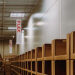 A warehouse interior with high shelves filled with boxes lining the aisle. "FLEX" banners hang from the ceiling. The atmosphere is organized and industrial.