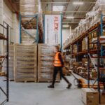 A person in a high-visibility vest moves quickly with a pallet jack in a neatly organized warehouse filled with boxes and pallets, conveying efficiency.