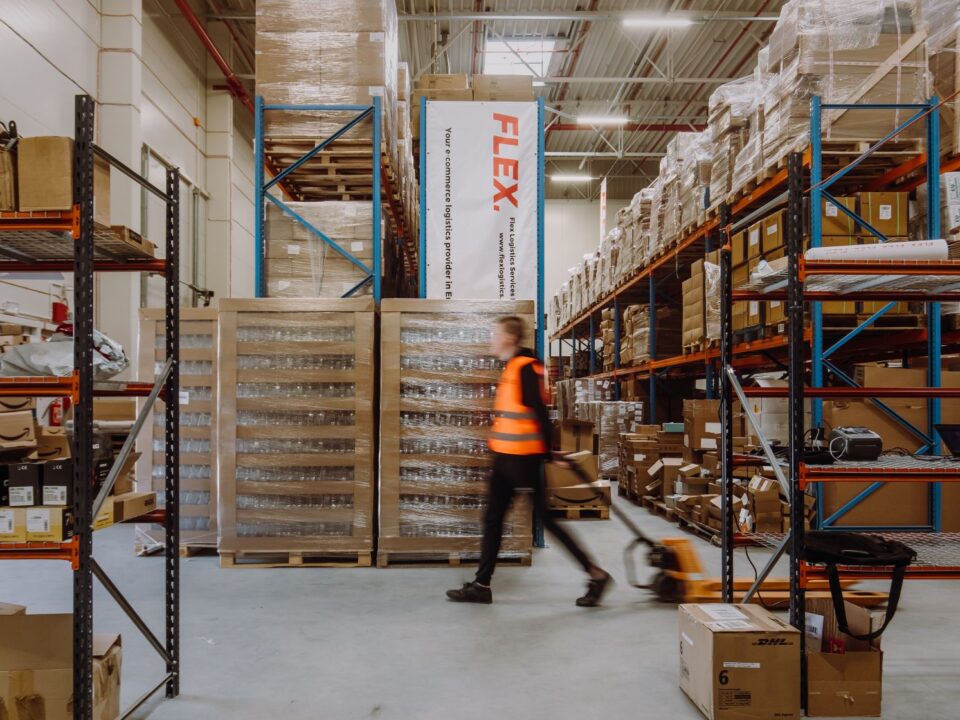 A person in a high-visibility vest moves quickly with a pallet jack in a neatly organized warehouse filled with boxes and pallets, conveying efficiency.