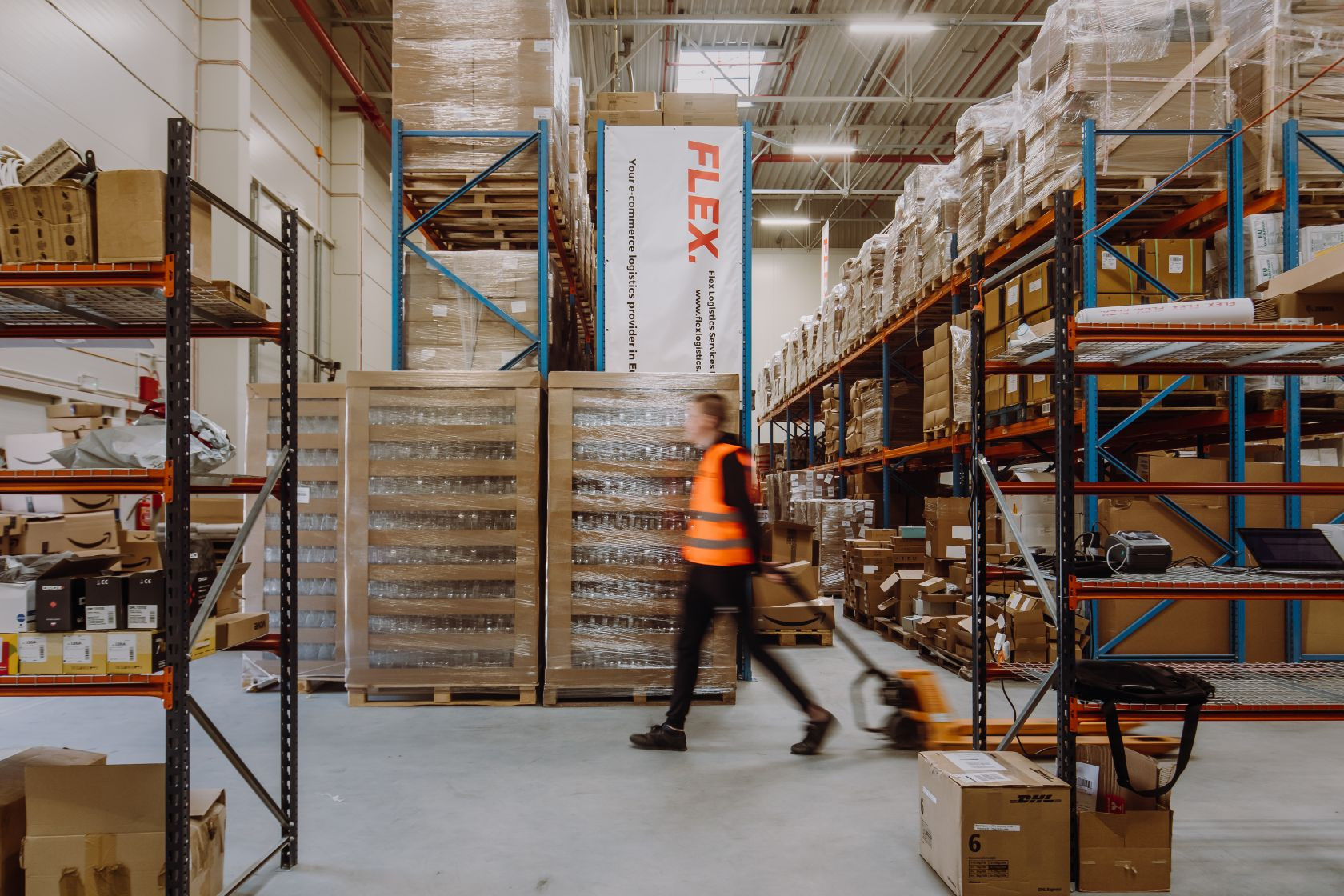A person in a high-visibility vest moves quickly with a pallet jack in a neatly organized warehouse filled with boxes and pallets, conveying efficiency.