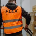 Worker in an orange "FLEX" safety vest operates a pallet jack in a warehouse filled with stacked boxes, suggesting efficient logistics work.