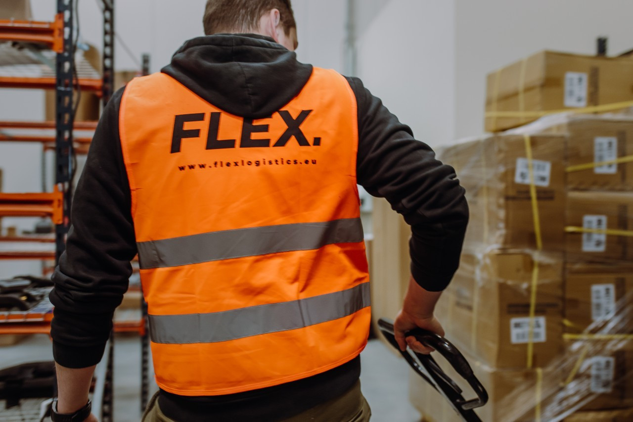 Worker in an orange "FLEX" safety vest operates a pallet jack in a warehouse filled with stacked boxes, suggesting efficient logistics work.