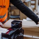 A worker in an orange vest labeled "FLEX" is handling a brown cardboard box. Nearby, black and white product boxes are stacked neatly. The setting appears to be a warehouse, conveying a sense of organization and efficiency.