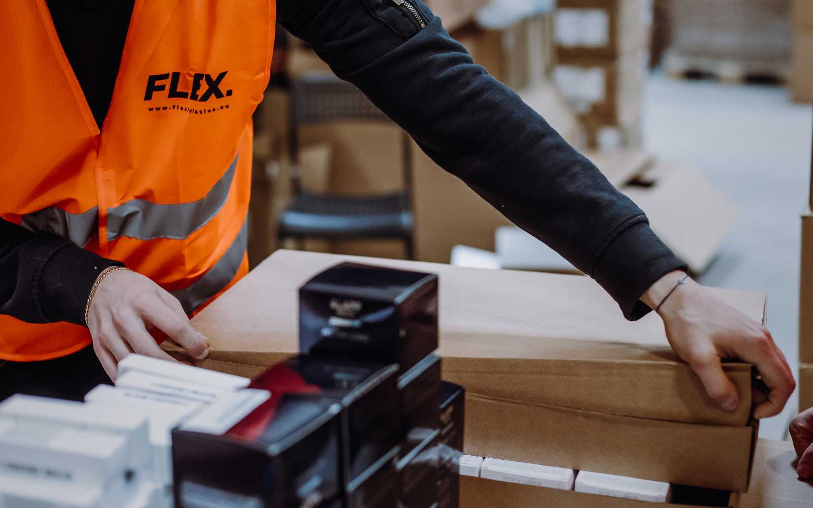 A worker in an orange vest labeled "FLEX" is handling a brown cardboard box. Nearby, black and white product boxes are stacked neatly. The setting appears to be a warehouse, conveying a sense of organization and efficiency.