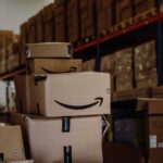 Stack of Amazon boxes in a warehouse. The setting includes shelves filled with cardboard packages, conveying organization and high activity.