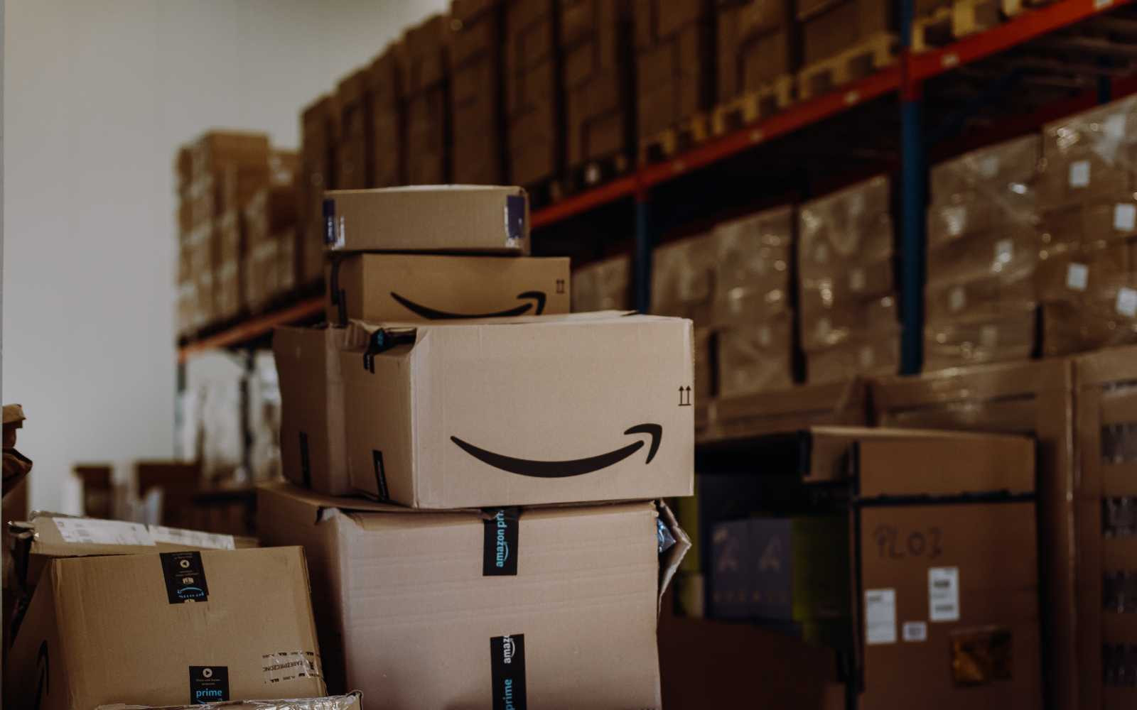 Stack of Amazon boxes in a warehouse. The setting includes shelves filled with cardboard packages, conveying organization and high activity.