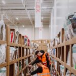 Two workers in orange vests organize shelves in a warehouse filled with products in cardboard boxes. Bright lighting and high ceiling create a busy atmosphere.