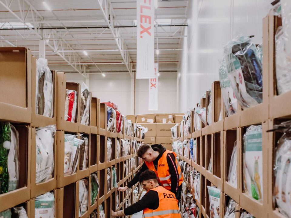 Two workers in orange vests organize shelves in a warehouse filled with products in cardboard boxes. Bright lighting and high ceiling create a busy atmosphere.