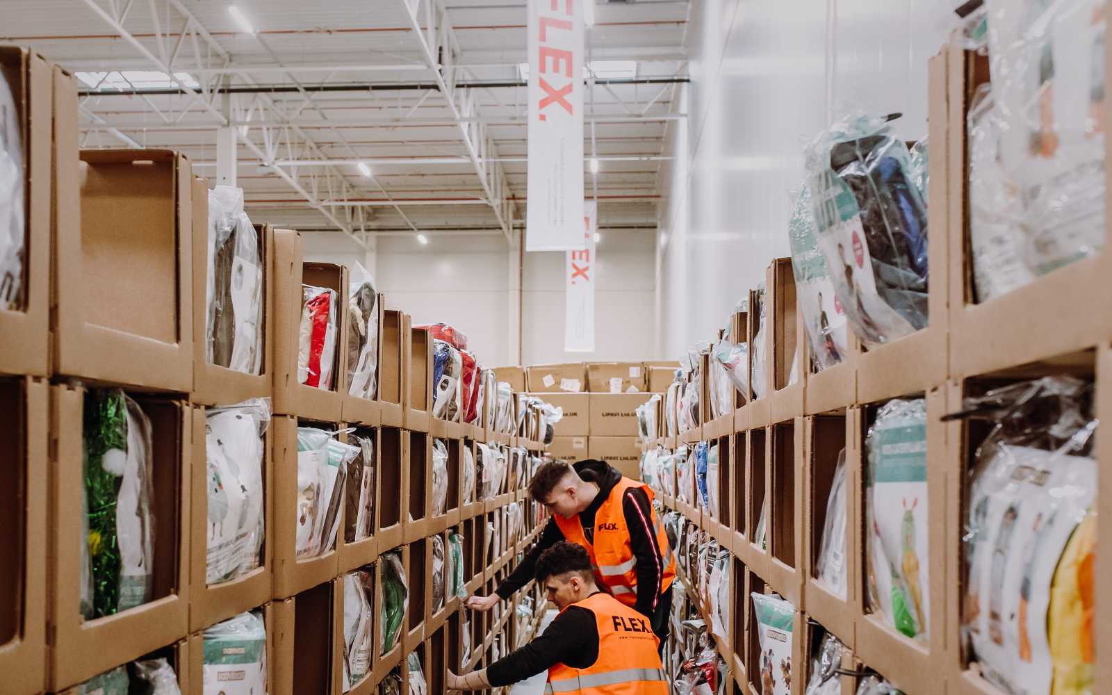 Two workers in orange vests organize shelves in a warehouse filled with products in cardboard boxes. Bright lighting and high ceiling create a busy atmosphere.