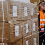 A worker in an orange safety vest operates a pallet jack, moving large wrapped boxes in a warehouse. The scene conveys focused activity and efficiency.