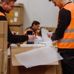 Workers in orange vests organize packages in a warehouse. Boxes labeled "Fragile" are stacked nearby. The atmosphere is focused and busy.