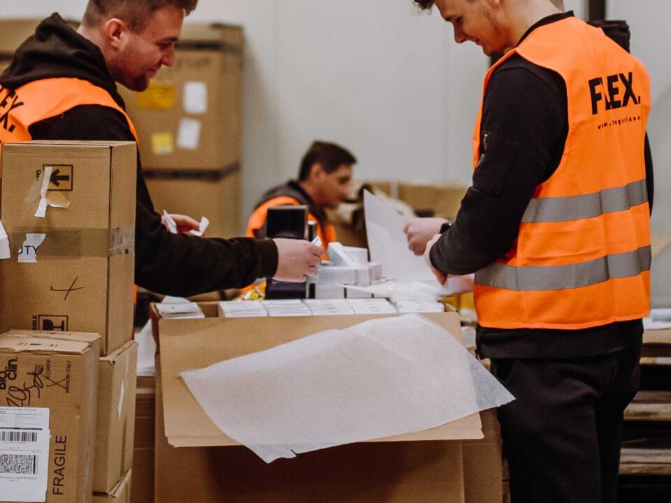Workers in orange vests organize packages in a warehouse. Boxes labeled "Fragile" are stacked nearby. The atmosphere is focused and busy.