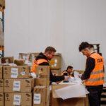Two workers in orange vests sort packages in a warehouse with boxes and shelves. The scene is busy, conveying productivity and organization.