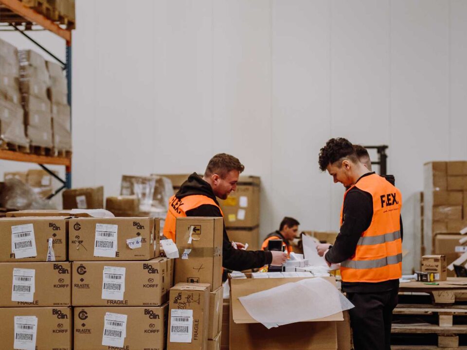 Two workers in orange vests sort packages in a warehouse with boxes and shelves. The scene is busy, conveying productivity and organization.