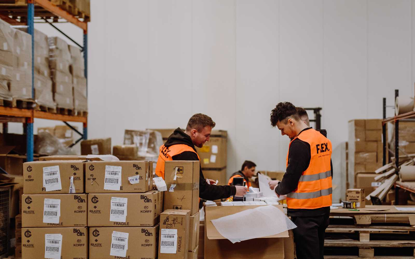 Two workers in orange vests sort packages in a warehouse with boxes and shelves. The scene is busy, conveying productivity and organization.