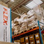 A warehouse scene with tall shelves holding stacked, wrapped pallets and cardboard boxes. A banner reads "FLEX" in bold red letters, suggesting logistics services.