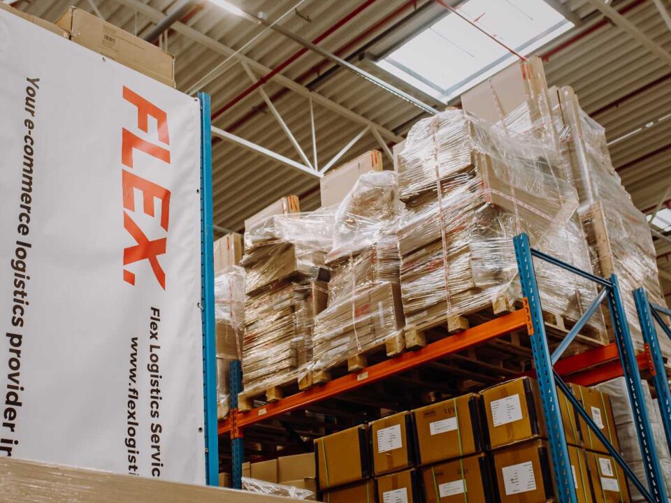 A warehouse scene with tall shelves holding stacked, wrapped pallets and cardboard boxes. A banner reads "FLEX" in bold red letters, suggesting logistics services.