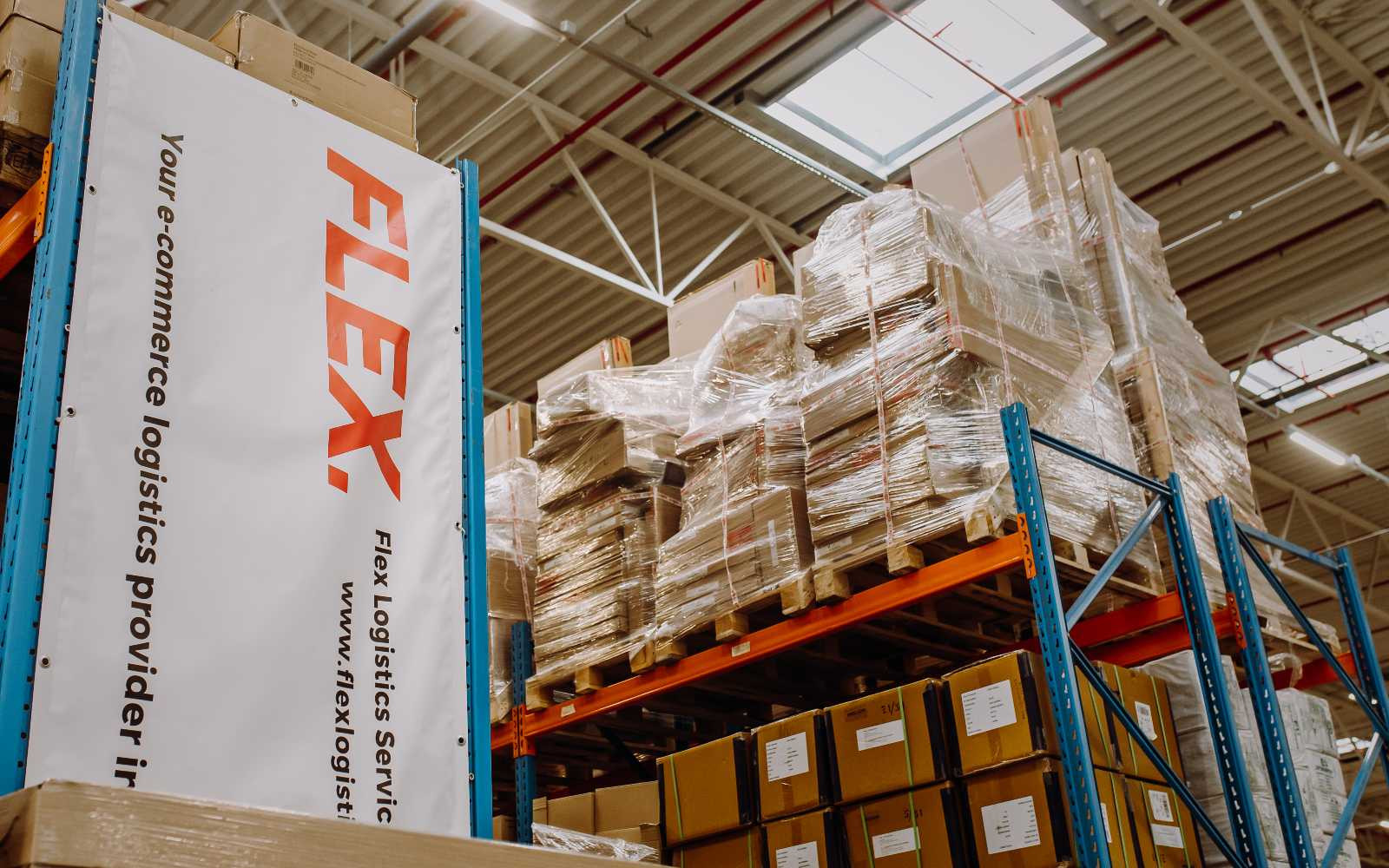 A warehouse scene with tall shelves holding stacked, wrapped pallets and cardboard boxes. A banner reads "FLEX" in bold red letters, suggesting logistics services.