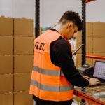 A warehouse worker in an orange vest uses a laptop on a metal rack. Boxes line shelves in the background, creating a busy, organized atmosphere.