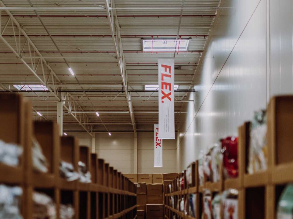 Spacious warehouse interior with tall shelves stacked with boxes. A banner reading “FLEX” hangs from the ceiling, conveying an organized, industrious atmosphere.