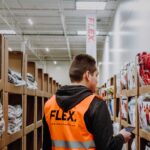 A worker in an orange vest marked "FLEX" is organizing items in a warehouse aisle lined with shelves of packaged goods, conveying efficiency and focus.