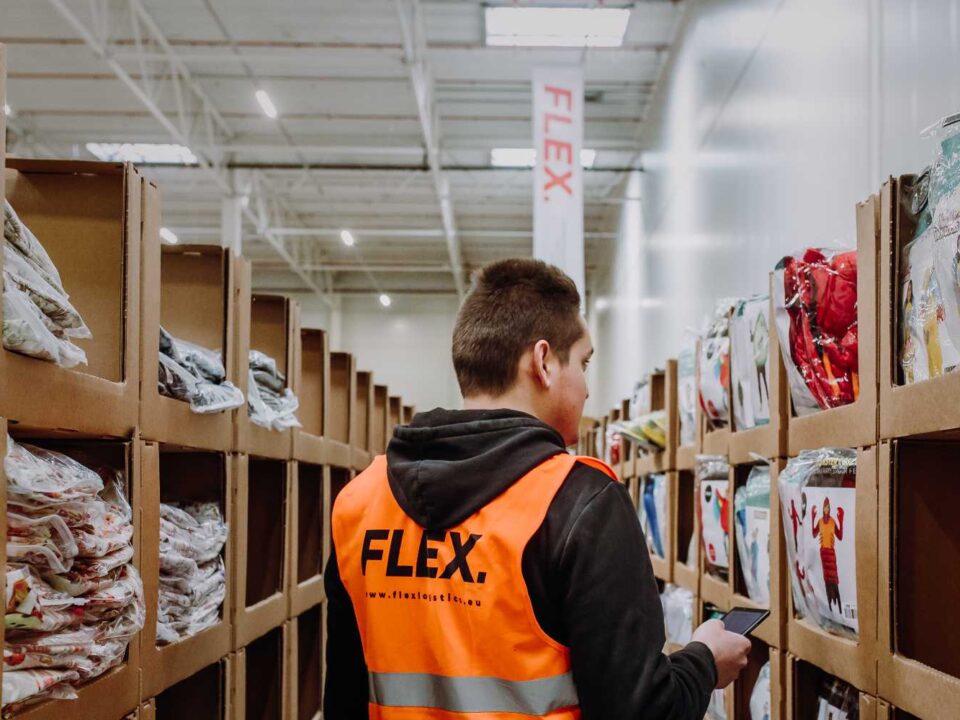 A worker in an orange vest marked "FLEX" is organizing items in a warehouse aisle lined with shelves of packaged goods, conveying efficiency and focus.