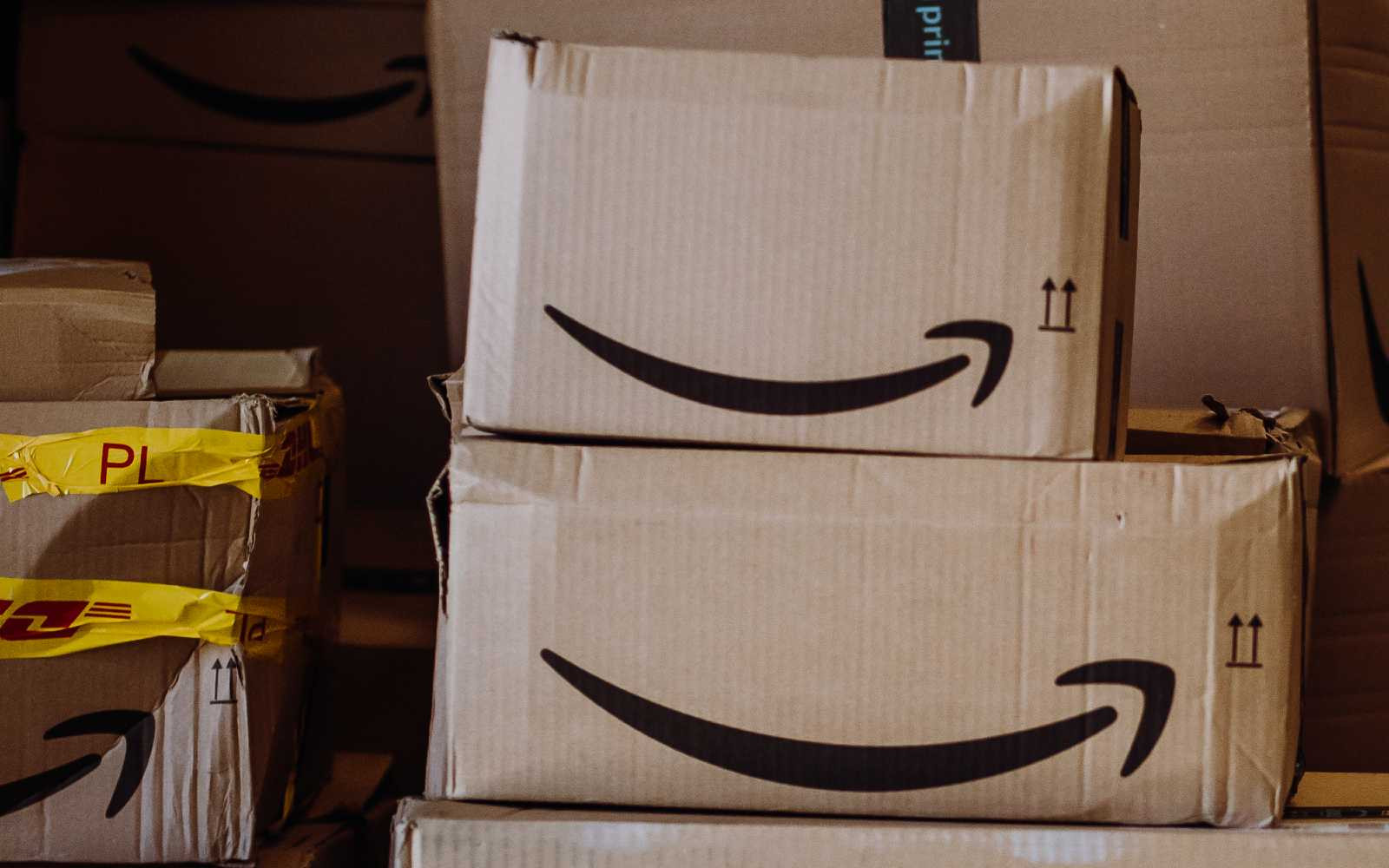 Stacked cardboard boxes with Amazon's smiling arrow logo and yellow tape, conveying a busy delivery or shipping environment.