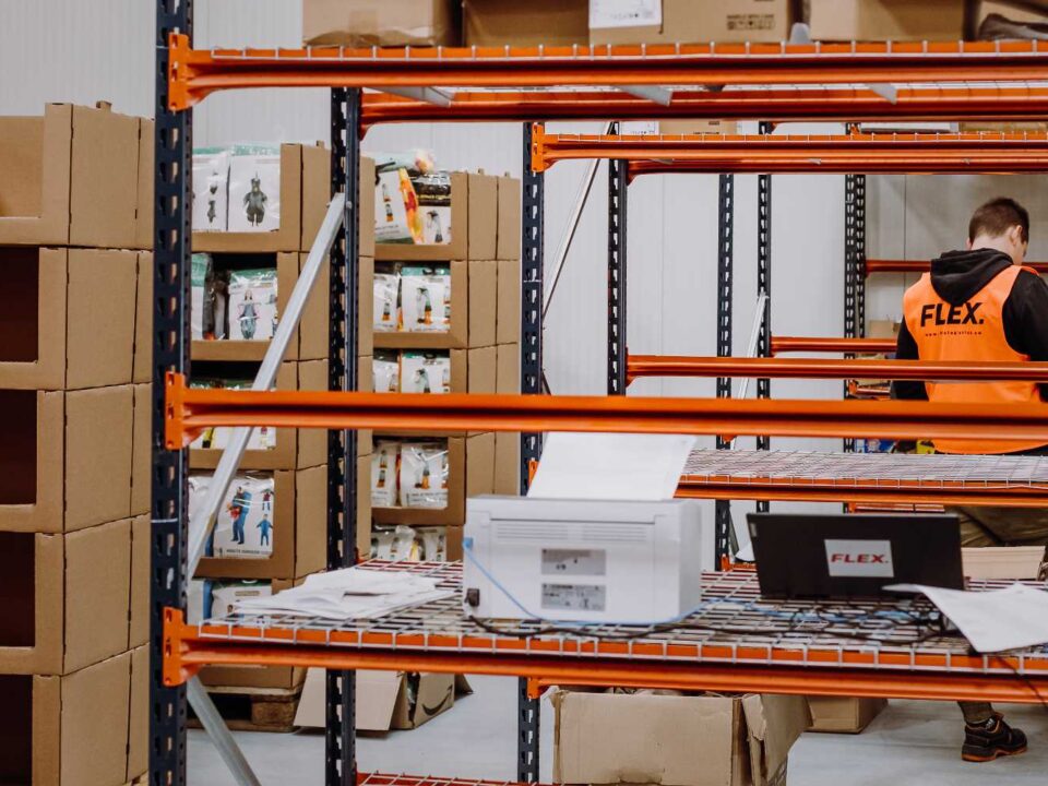 Warehouse scene showing empty orange metal shelves in a room with stacked cardboard boxes in the background. A man in a "FLEX" vest writes notes.