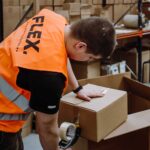 A person wearing an orange safety vest labeled "FLEX" handles a cardboard box in a warehouse. Shelves with more boxes are visible in the background. The tone is busy and industrial.