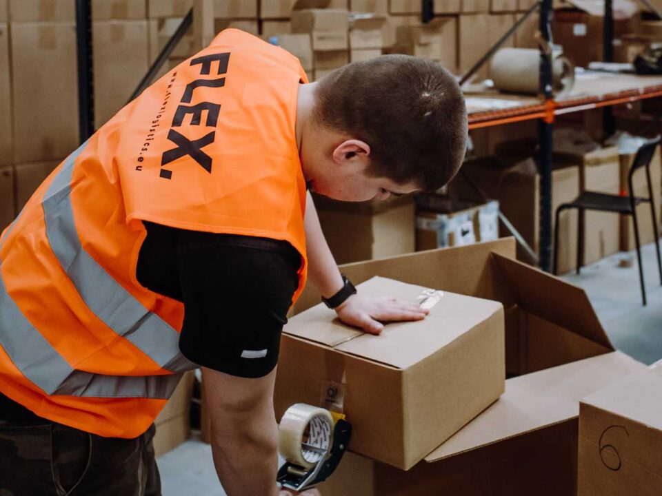 A person wearing an orange safety vest labeled "FLEX" handles a cardboard box in a warehouse. Shelves with more boxes are visible in the background. The tone is busy and industrial.
