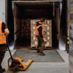 Two workers in orange safety vests load a pallet into a truck using a manual pallet jack in a warehouse setting. The scene is industrious and focused.