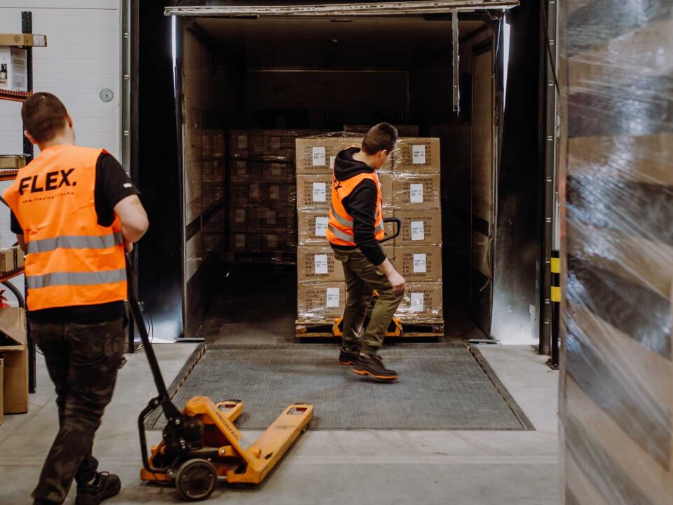 Two workers in orange safety vests load a pallet into a truck using a manual pallet jack in a warehouse setting. The scene is industrious and focused.