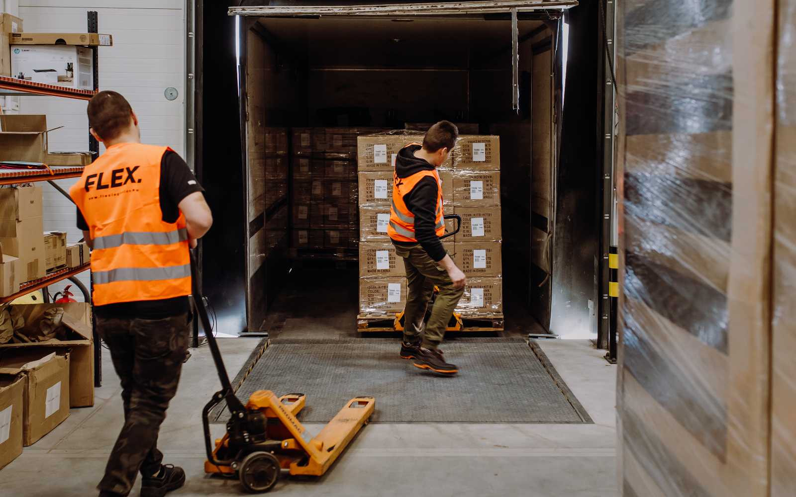 Two workers in orange safety vests load a pallet into a truck using a manual pallet jack in a warehouse setting. The scene is industrious and focused.