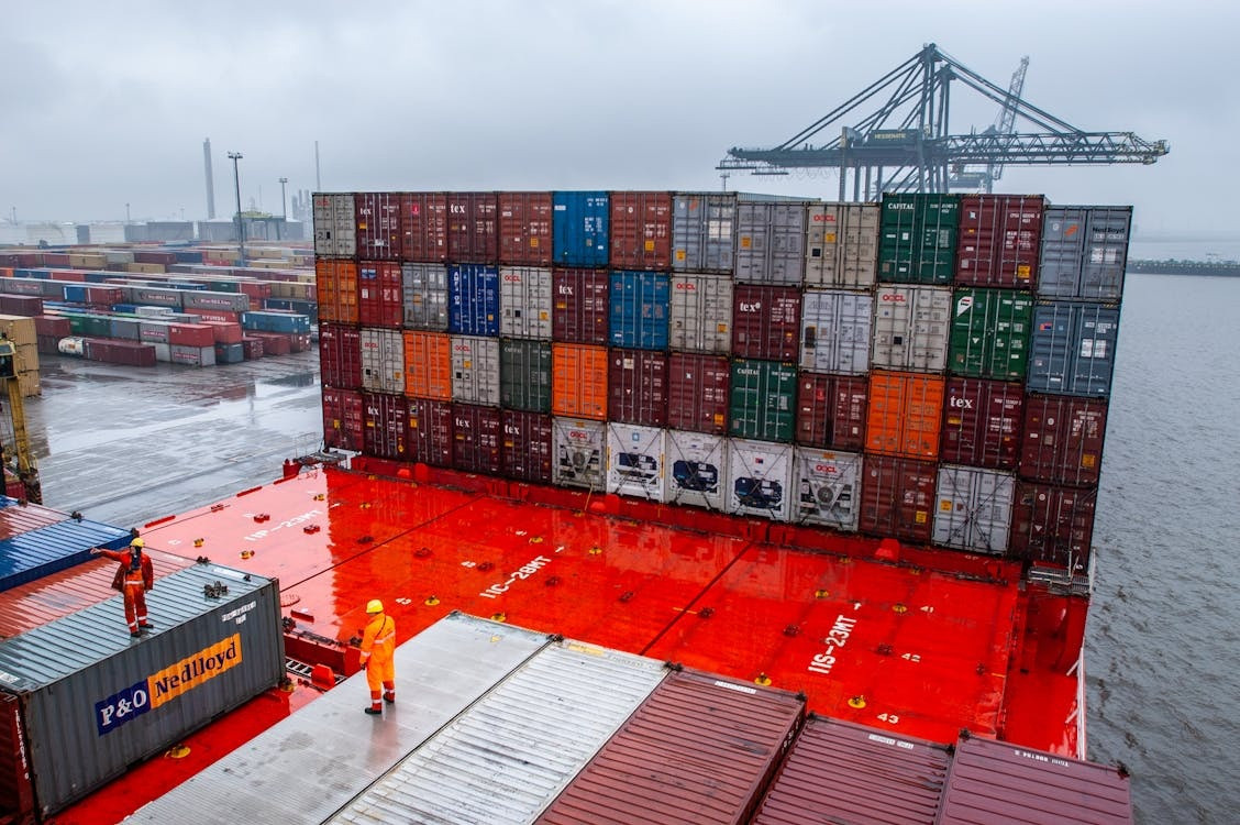 Stacked shipping containers on a vibrant red deck in a busy port. Dockworkers in orange gear attend to containers under overcast skies. Industrial and bustling.