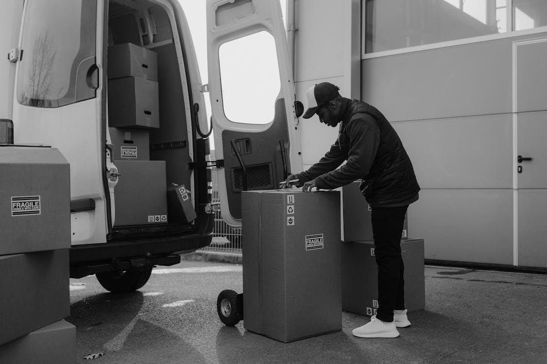 Man loading cardboard boxes labeled "Fragile" into a van on a dolly. He wears a cap and jacket, focused on work. It's a grayscale image.