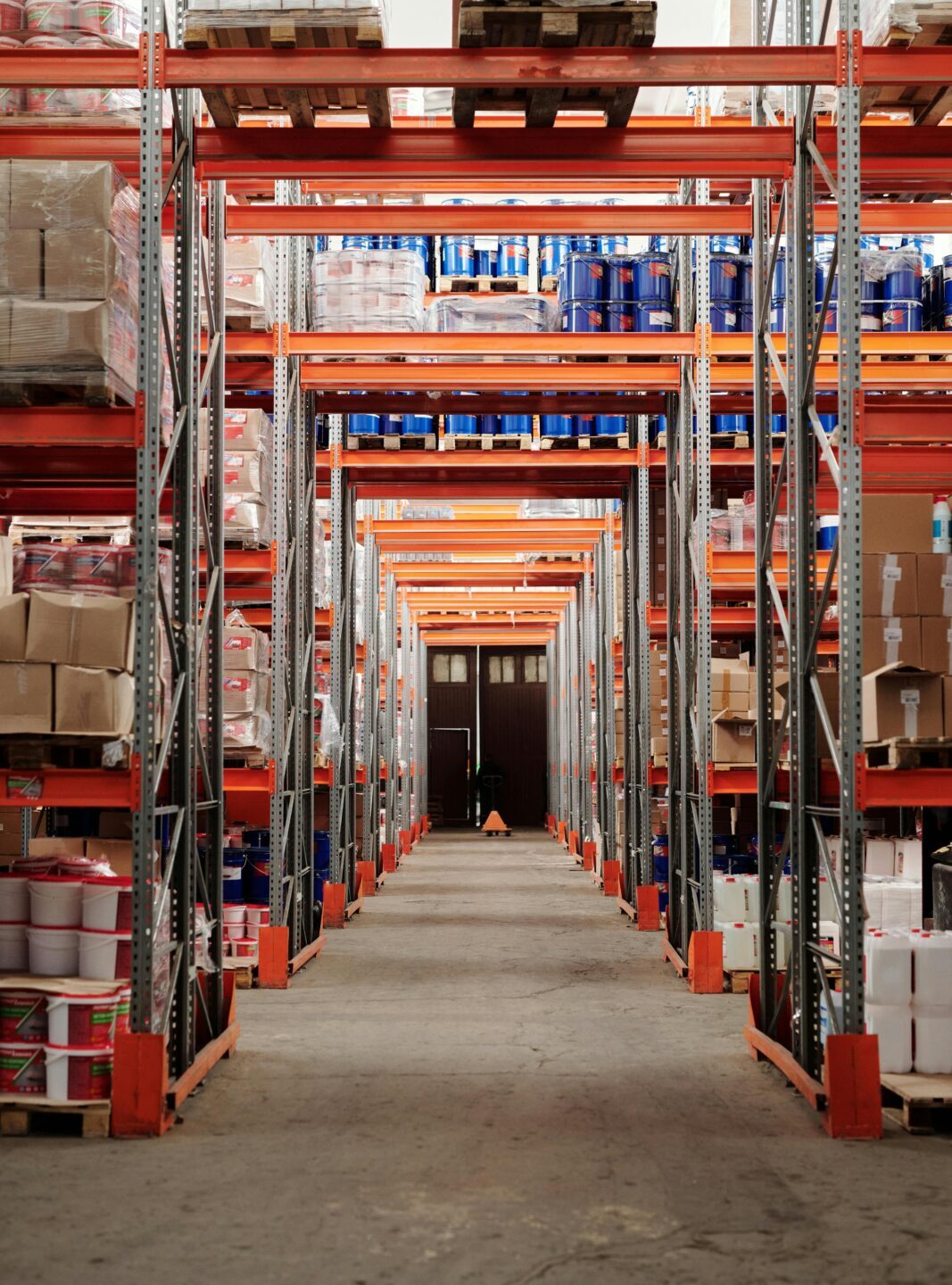 A warehouse aisle with tall metal shelves stacked with various boxes and products. The shelves create a symmetrical, organized pattern. The atmosphere is industrial.