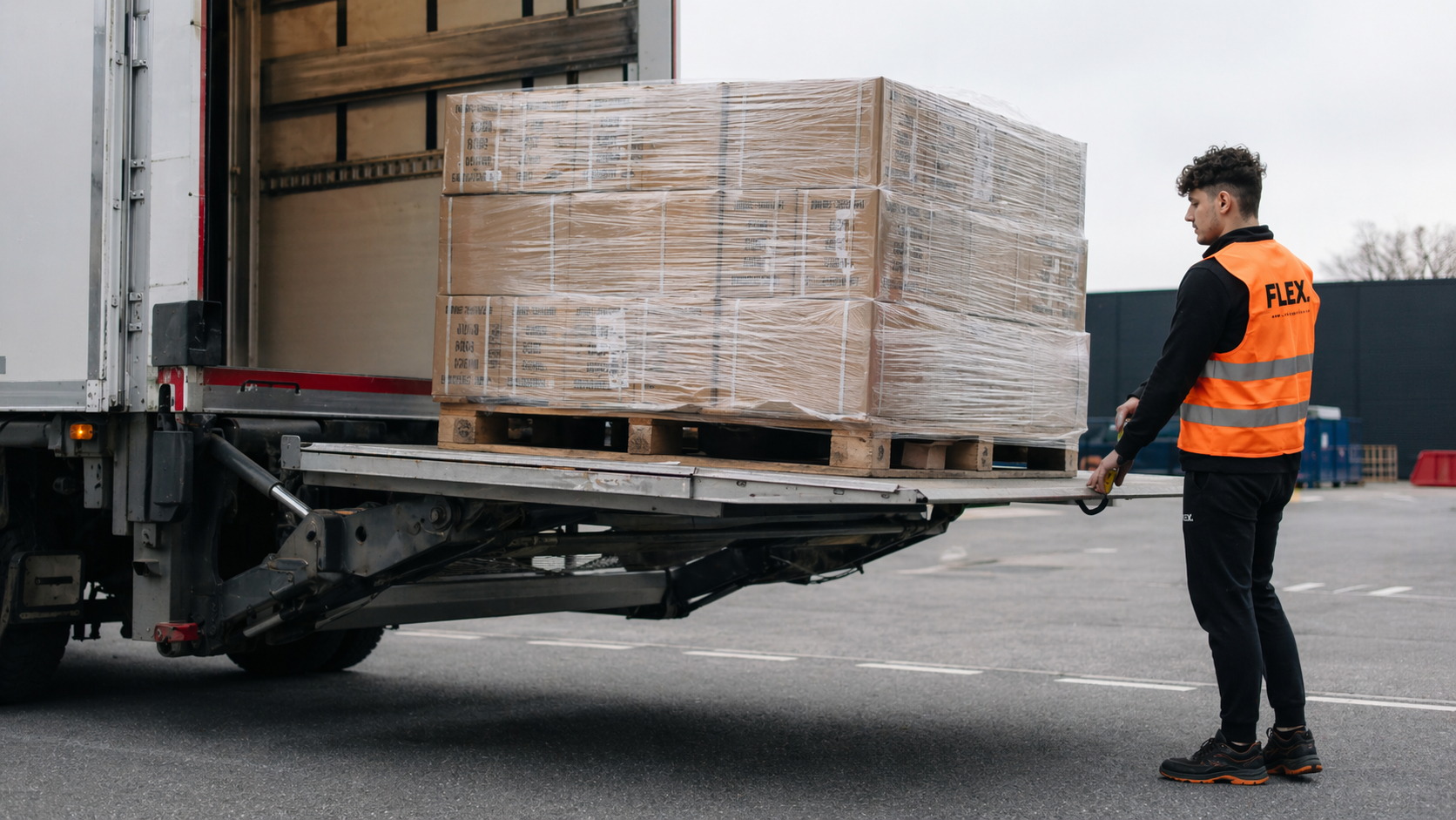 A stretch-wrapped pallet of boxes descending on a truck tail-lift to the tarmac, with a FLEX. worker guiding the operation