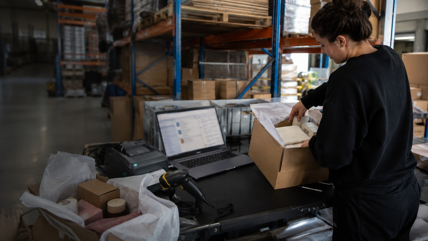 A warehouse operator packs mixed online store orders at a fulfillment station with open cartons, a scanner, a label printer, and a blurred order dashboard nearby.